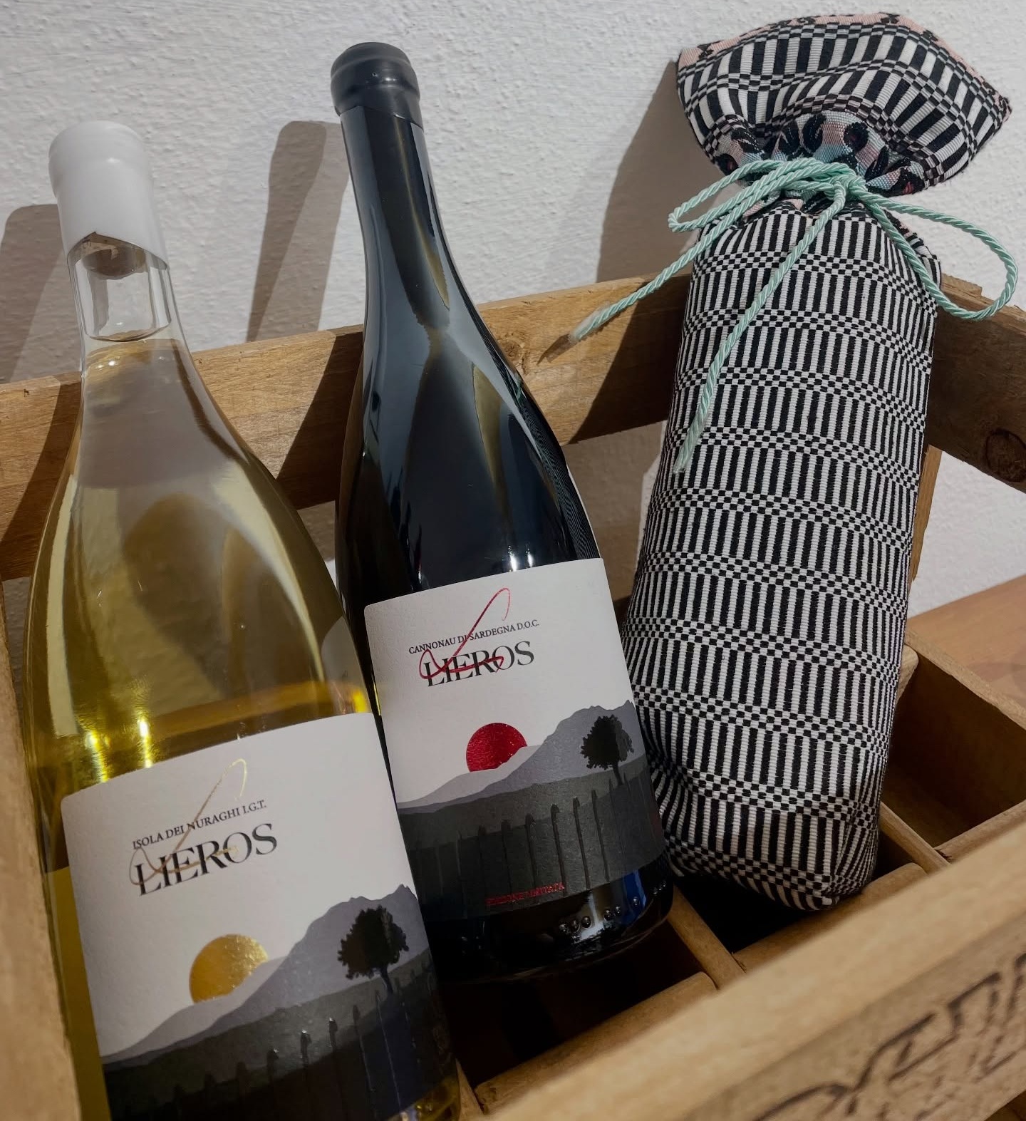 Bottiglie di vino bianco e Cannonau di Sardegna DOC Lieros in una cassetta di legno accanto a una confezione in tessuto